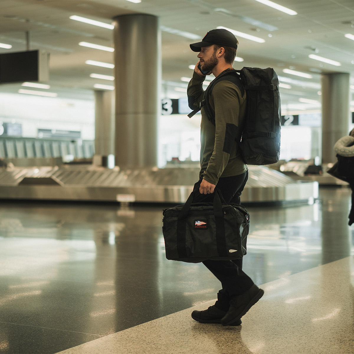 Man walking in airport with backpack and duffel bag, talking on phone near baggage claim area.