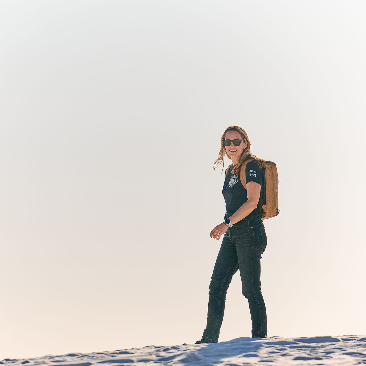 Woman with sunglasses and a backpack standing on a sandy surface under a bright, clear sky.