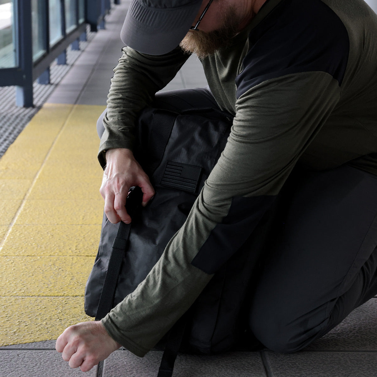 Man kneeling, opening a black backpack on a tiled floor near a yellow tactile paving strip.