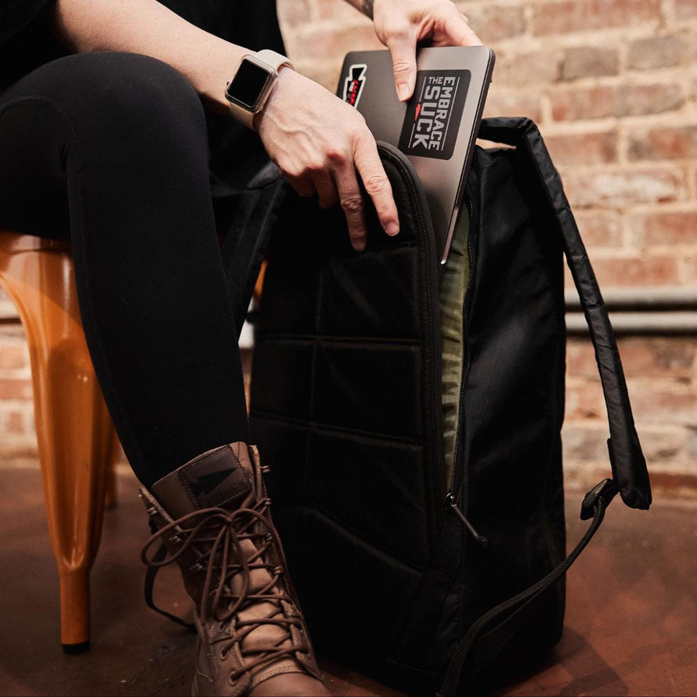 A person places a laptop into a black backpack while seated on a wooden chair by a brick wall.