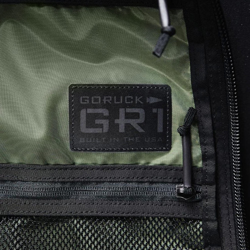 A close-up of a GORUCK GR1 label on green fabric inside a backpack, with black mesh and zippers nearby.