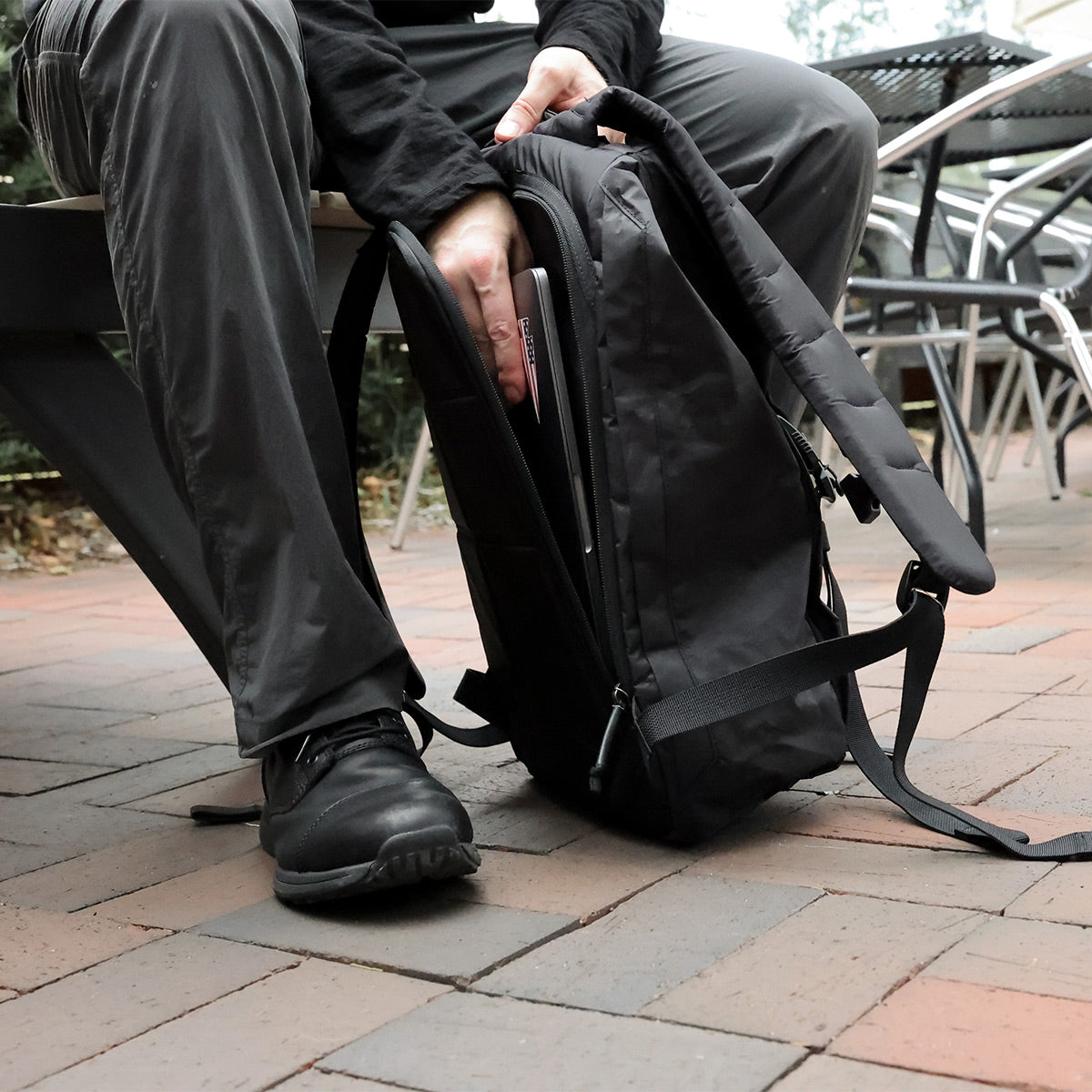 Person sitting outdoors, placing a laptop inside a black backpack on a brick-paved surface.