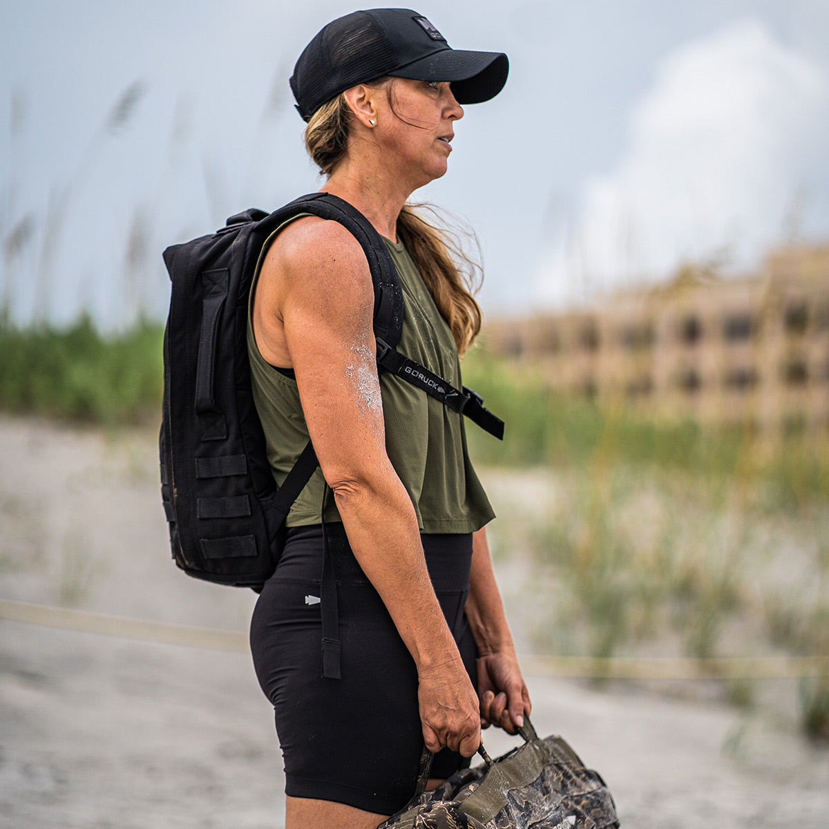 Woman in workout gear with a backpack and cap stands outdoors holding a heavy bag, with greenery in the background.