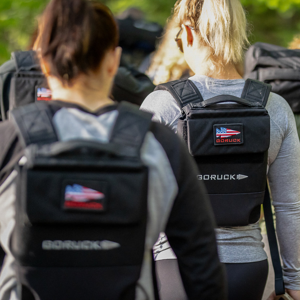 Two people wearing backpacks with GORUCK logos, walking outdoors in athletic clothing.