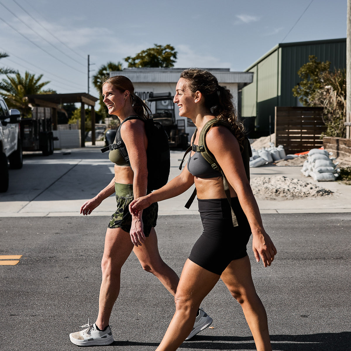 Two women in athletic wear and backpacks walk and smile on a sunny street.