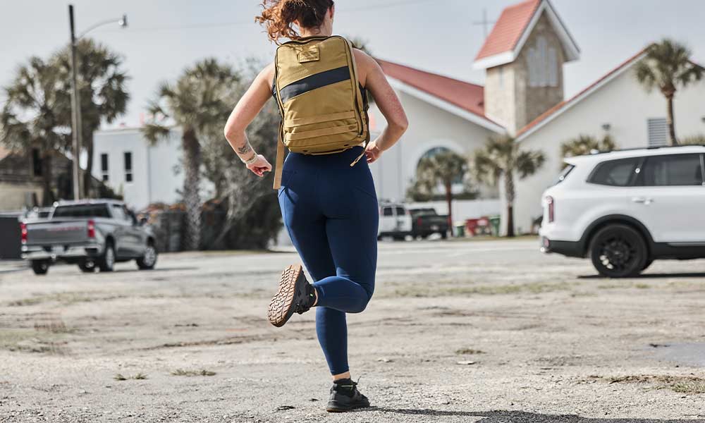 A person in athletic wear runs outdoors on a gravel lot, with buildings and parked cars in the background.