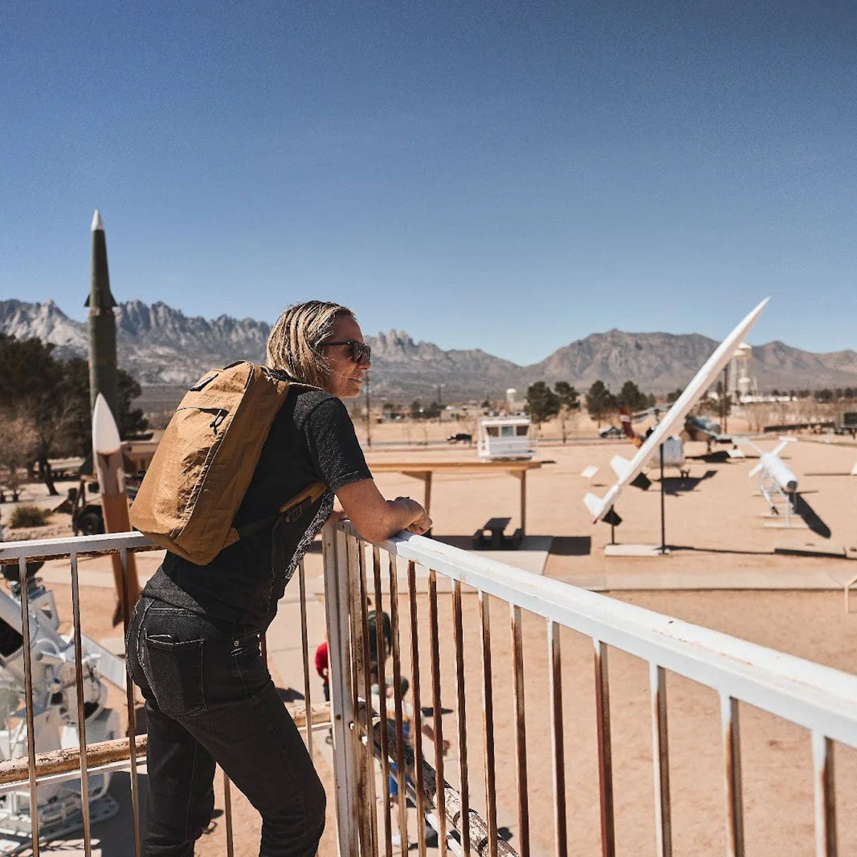 A person with a backpack leans on a railing, overlooking a desert museum with rockets and mountains in the background.