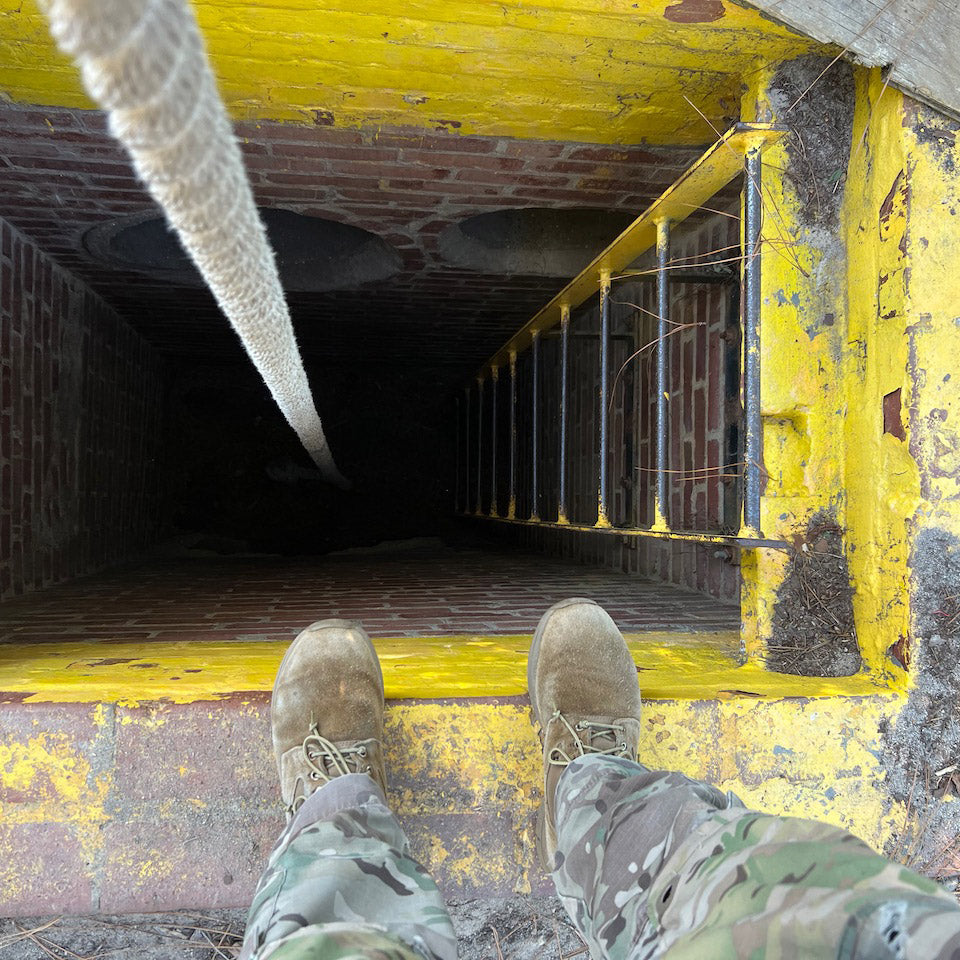 A person in camouflage pants and boots stands at the edge of a deep, brick-lined hole with a yellow ladder.