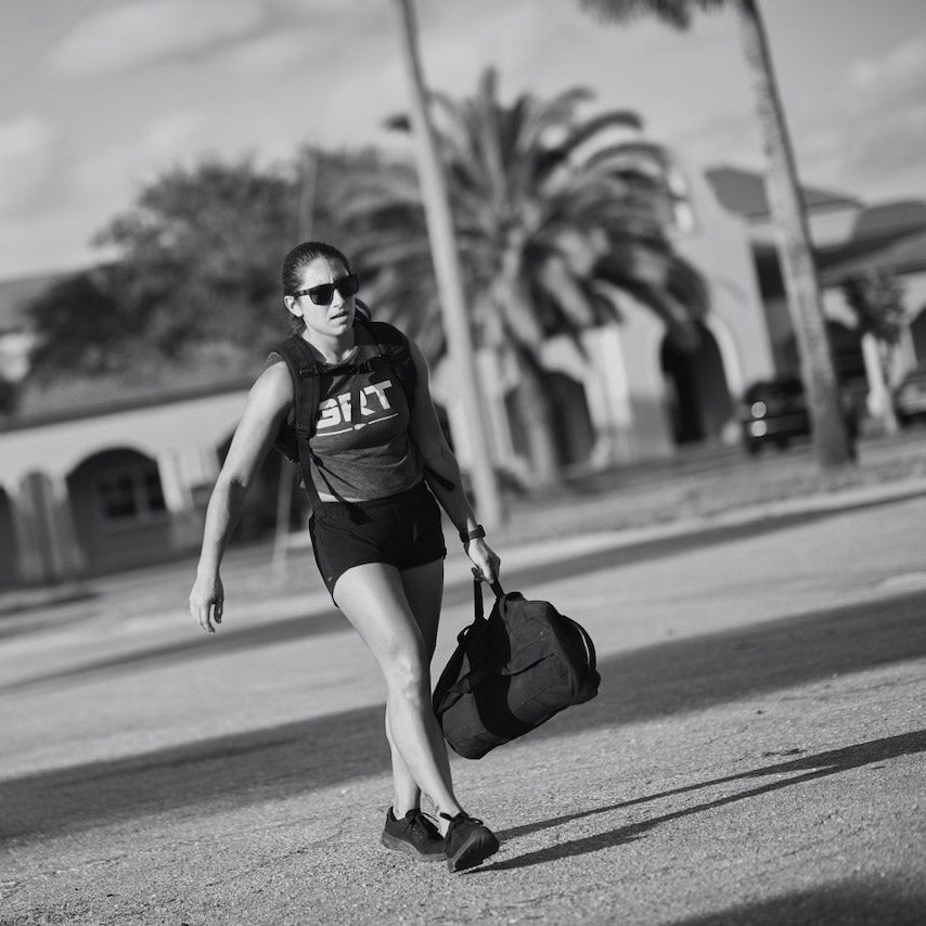 A woman in athletic wear walks outdoors with a gym bag, palm trees and buildings in the background. Black and white.