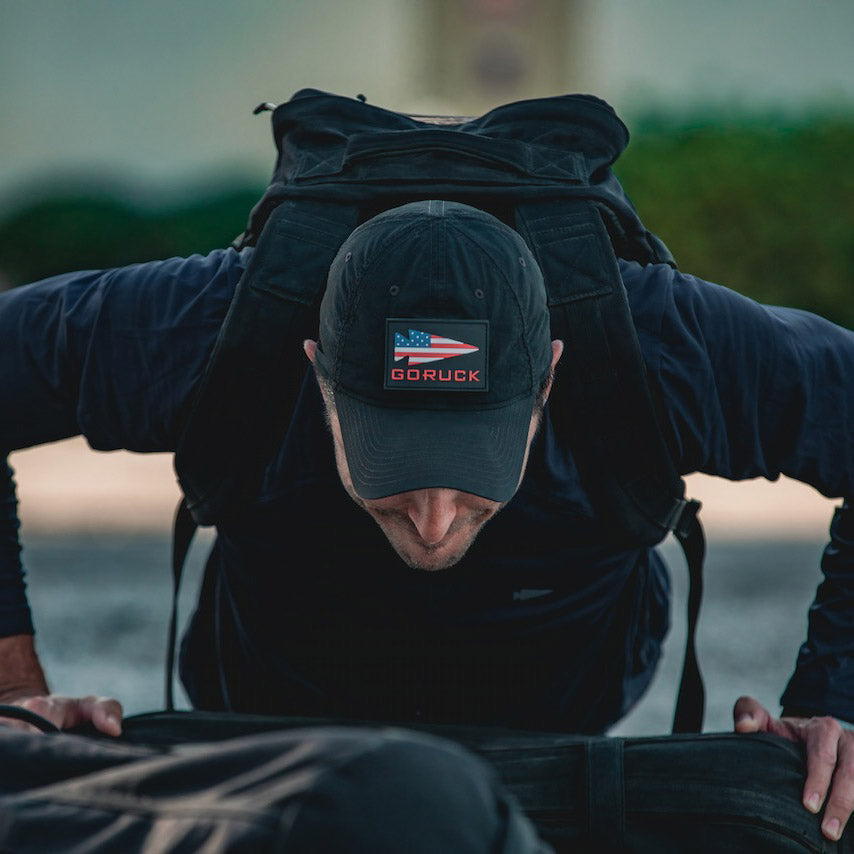 Person wearing a black GORUCK cap and backpack doing push-ups outdoors.