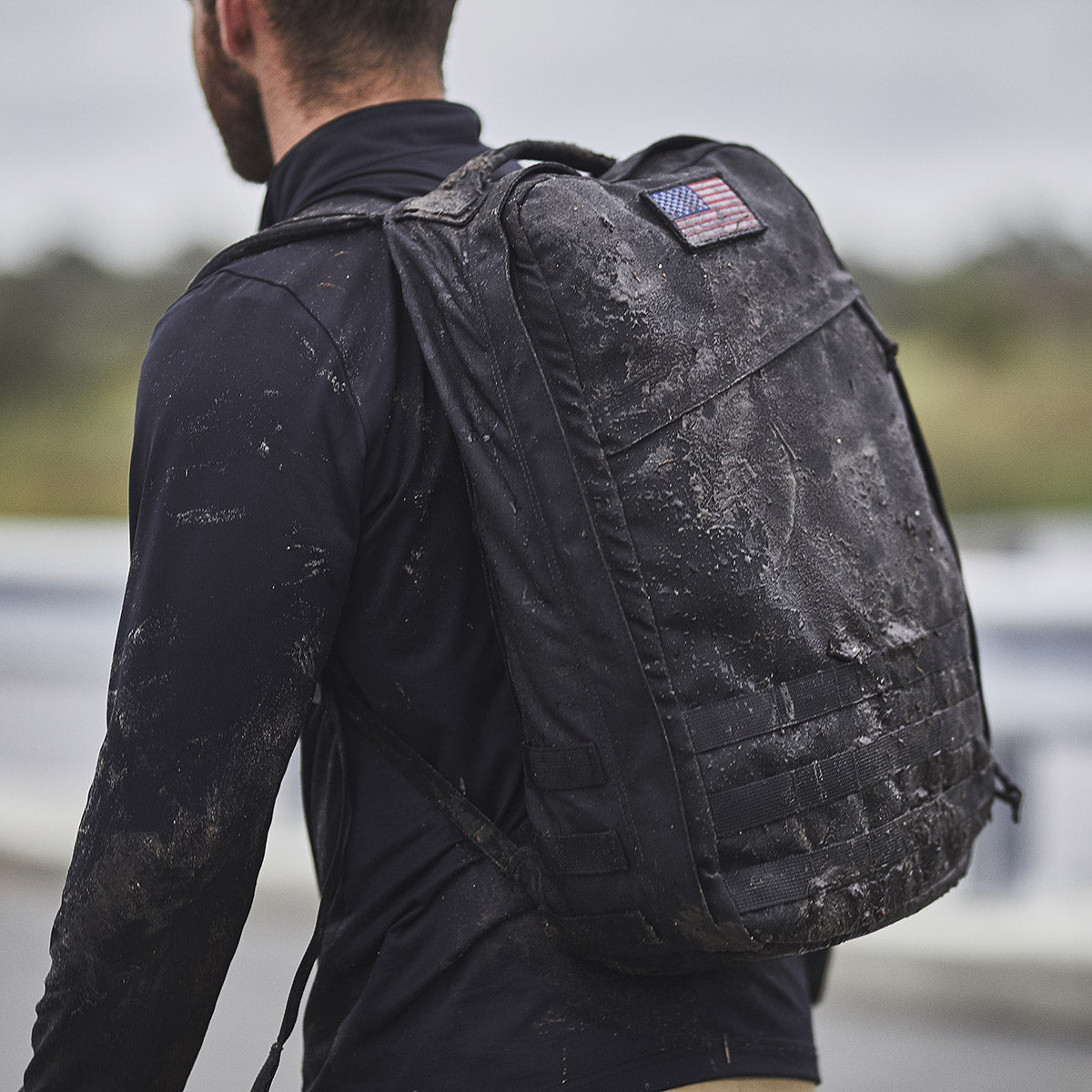 A person wearing a muddy black backpack with a U.S. flag patch, standing outdoors.