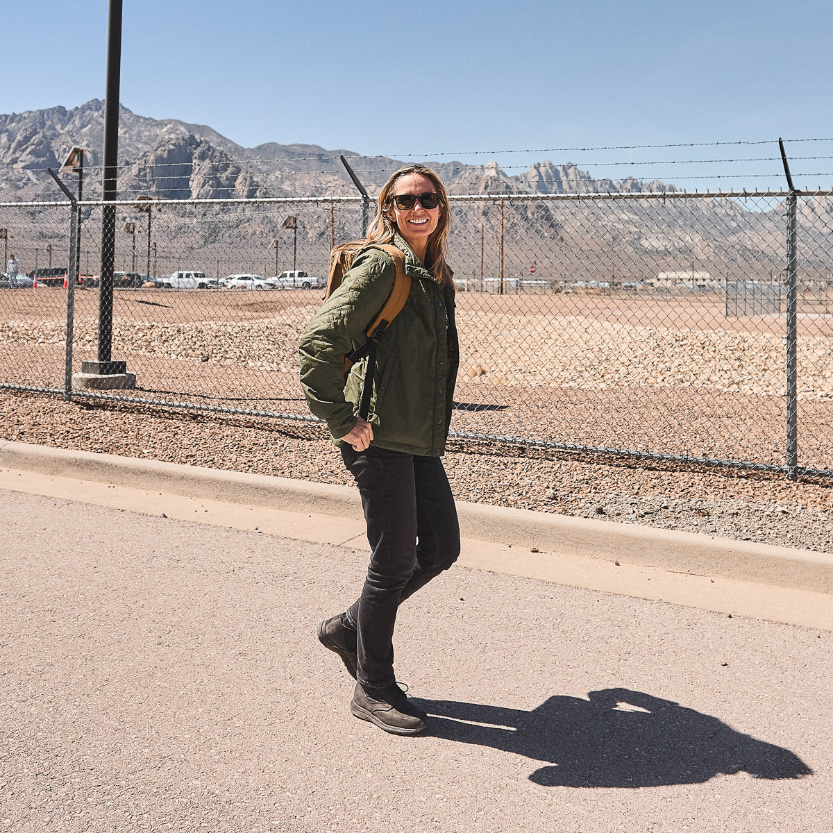 Woman wearing sunglasses and a green jacket smiles while walking near a fence with mountains in the background.