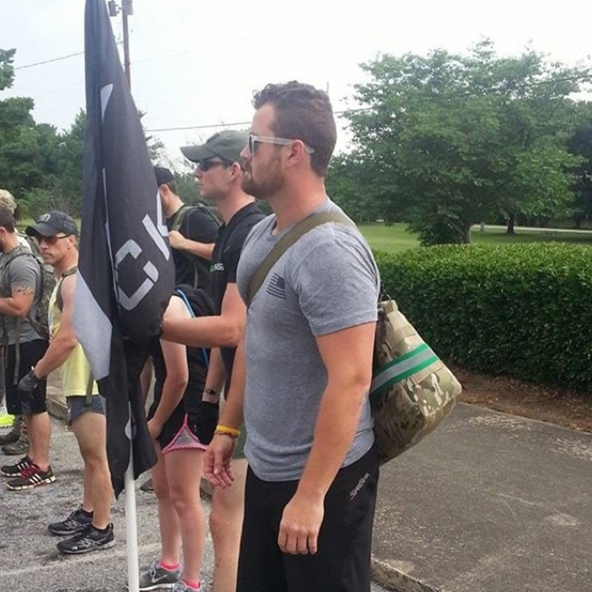 A group of people stand outdoors near a flag, some wearing athletic clothing and carrying bags.