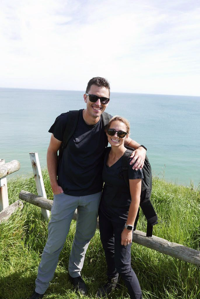 Two people in sunglasses stand smiling on a grassy cliff overlooking the ocean on a sunny day.