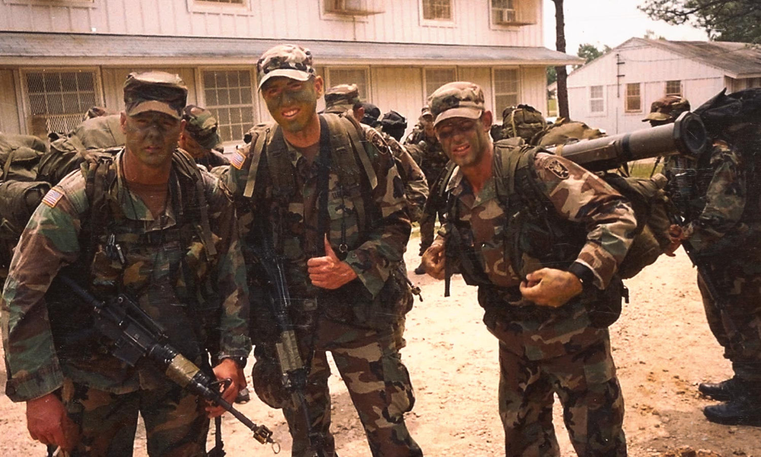 Three soldiers in camouflage uniforms and face paint smile for the camera with gear and weapons outdoors.
