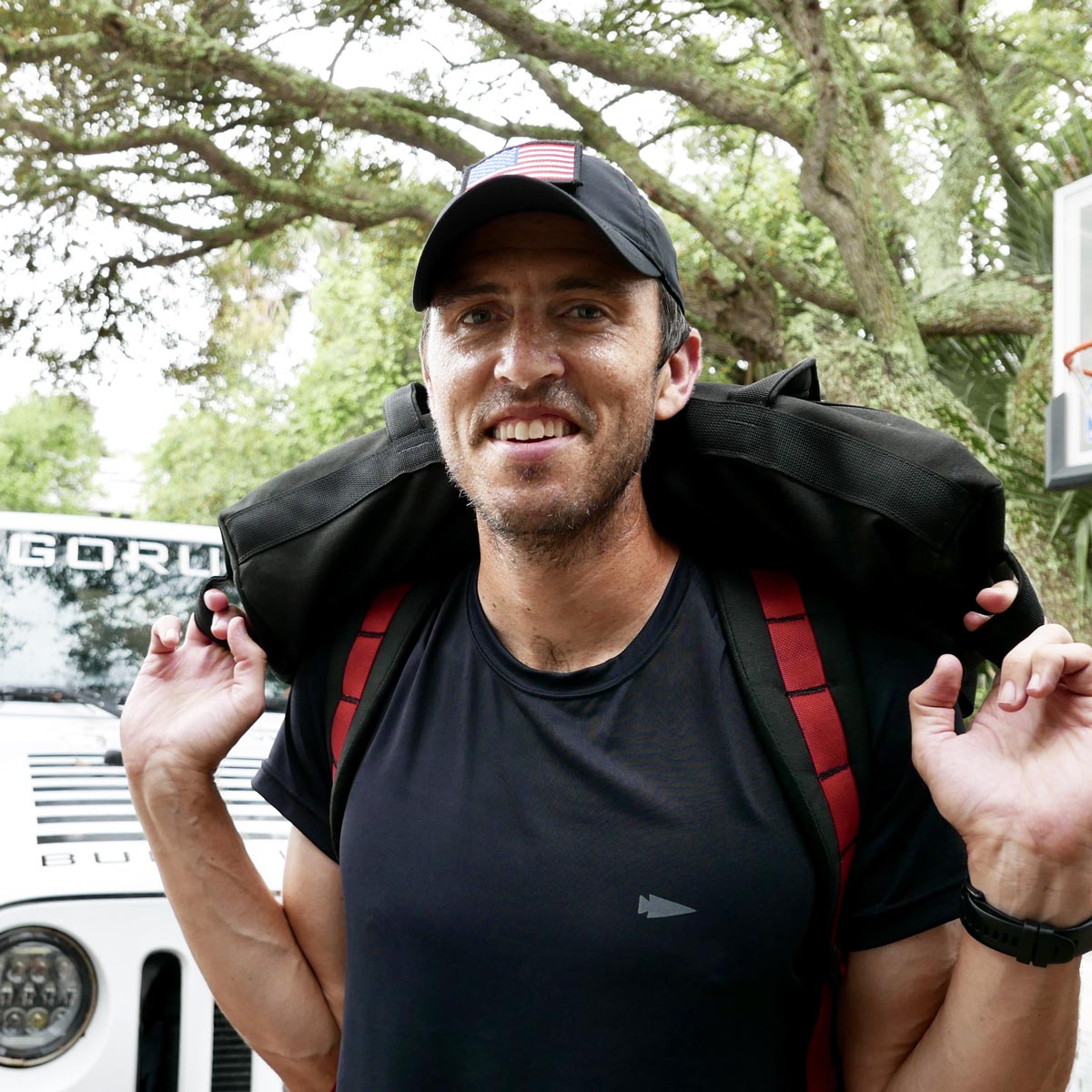 Man in a black shirt and cap smiles, carrying a black bag over his shoulders outdoors near a white vehicle.