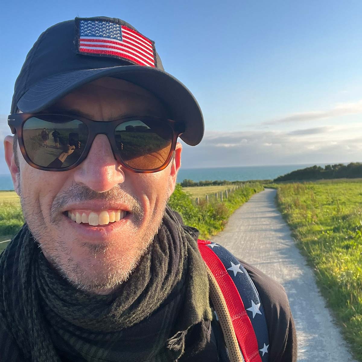 Smiling man in sunglasses and cap with US flag patch, standing on a sunny rural path with green fields.