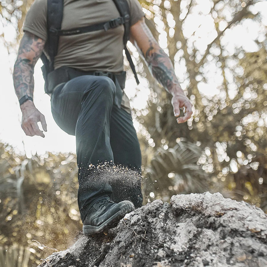 A person with tattoos hikes outdoors, kicking up dust while stepping onto a rocky surface.