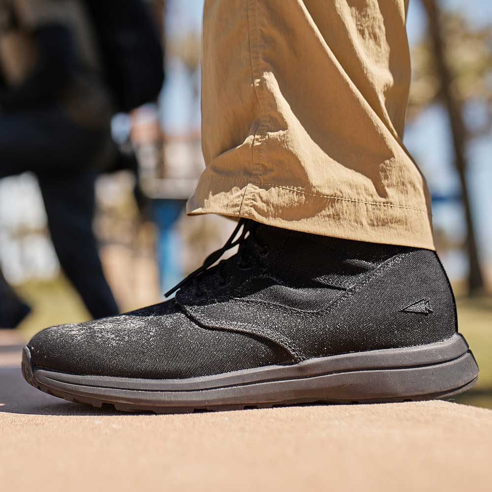 A person wearing black sneakers and beige pants standing outdoors on a sidewalk.