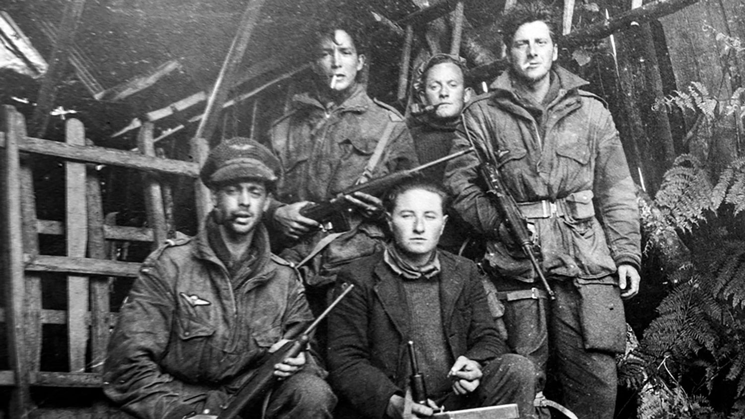 Five people in rugged clothing hold rifles, posing outdoors in front of a rustic wooden structure.