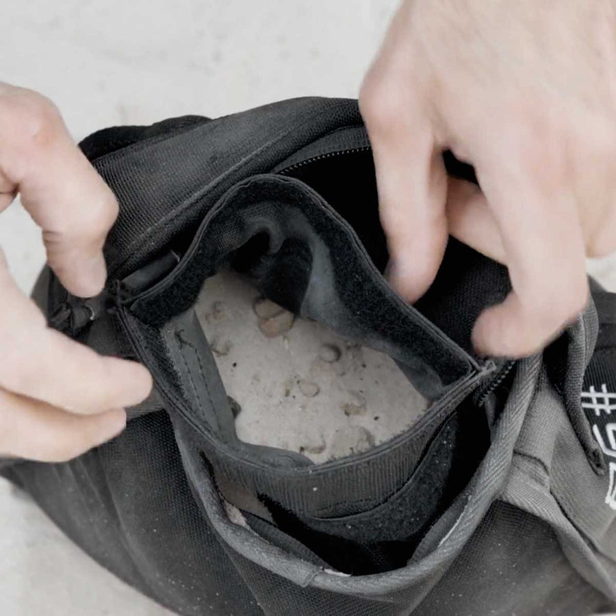 Hands opening a black bag filled with sand and small rocks on a light surface.