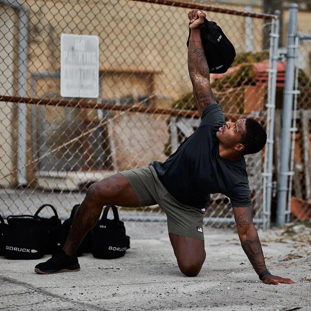 A man kneels on the ground, lifting a weighted bag overhead in an outdoor workout by a fence.