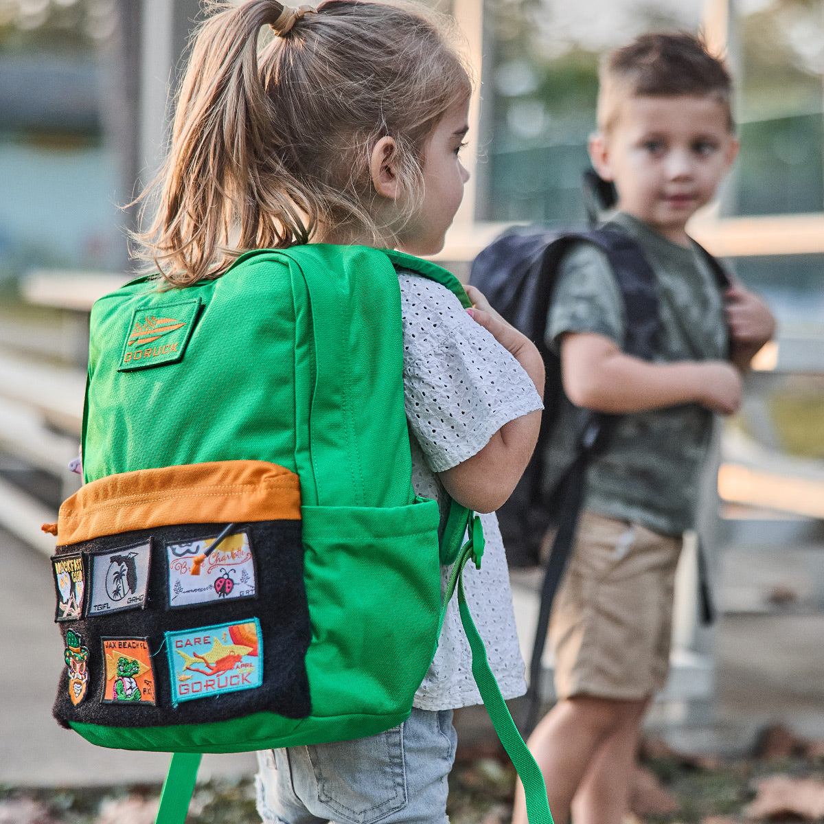 Young girl with a green backpack faces a boy with a dark backpack outdoors, both wearing casual clothes.