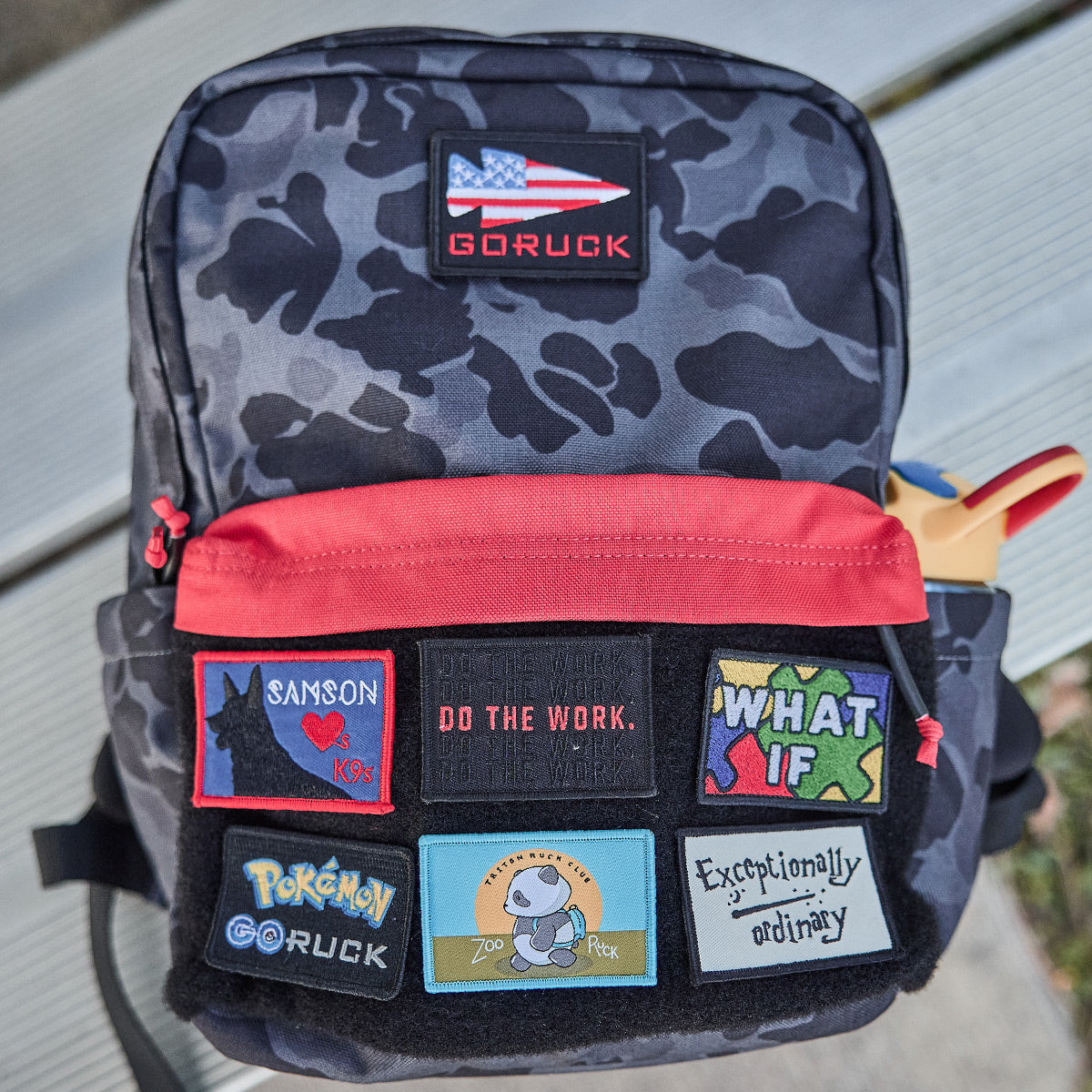 A black camo backpack with colorful patches on the front pocket, including Pokémon, Snoopy, and various slogans.