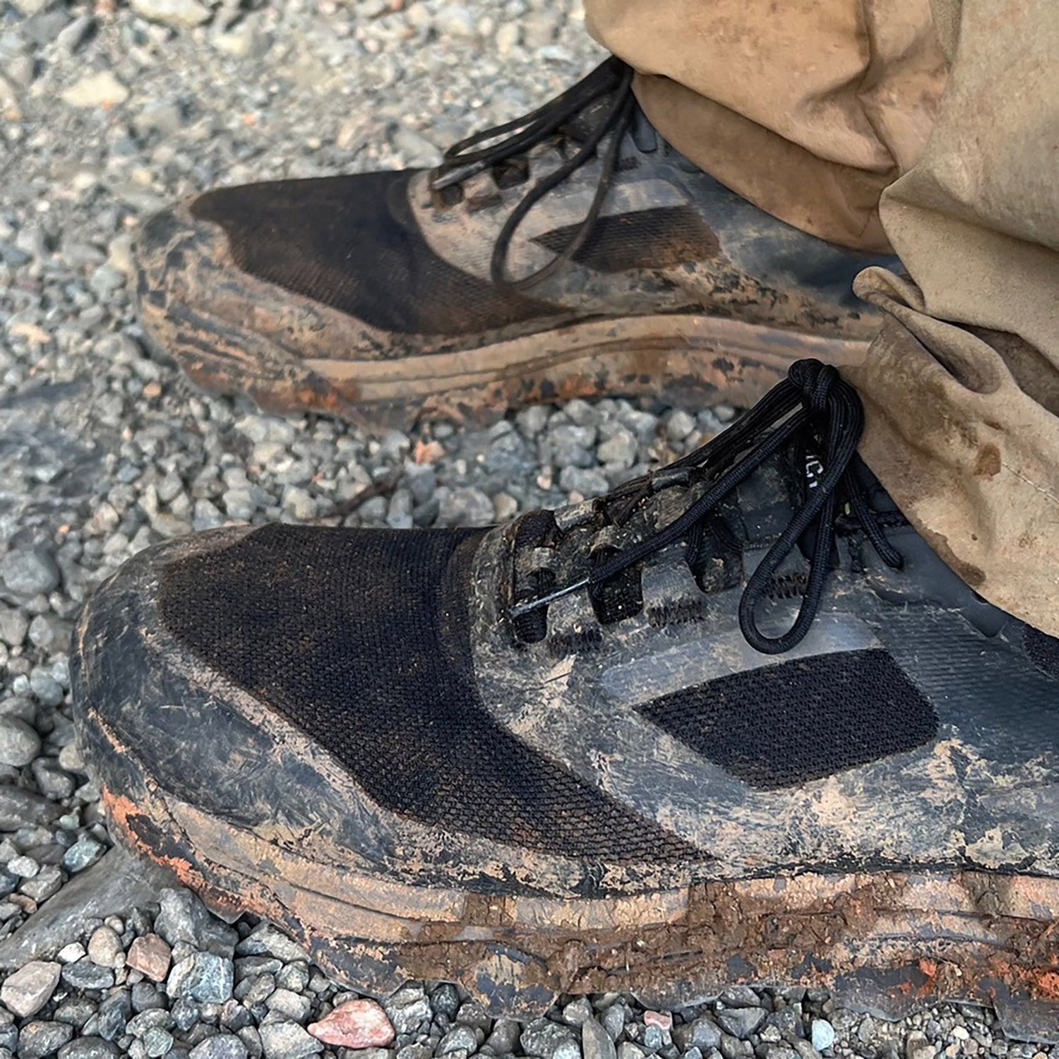 Close-up of muddy black sneakers and tan pants standing on gravel.