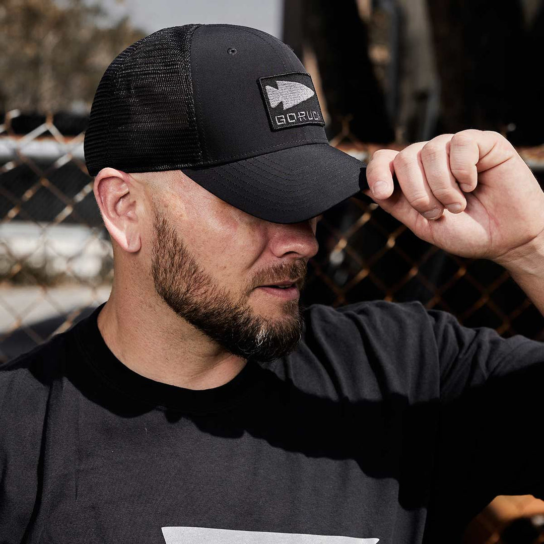 A man sporting a sweat-wicking black t-shirt and the GORUCK Performance Trucker Hat - GORUCK Spearhead, featuring an adjustable Velcro strap, adjusts his cap outdoors near a chain-link fence.