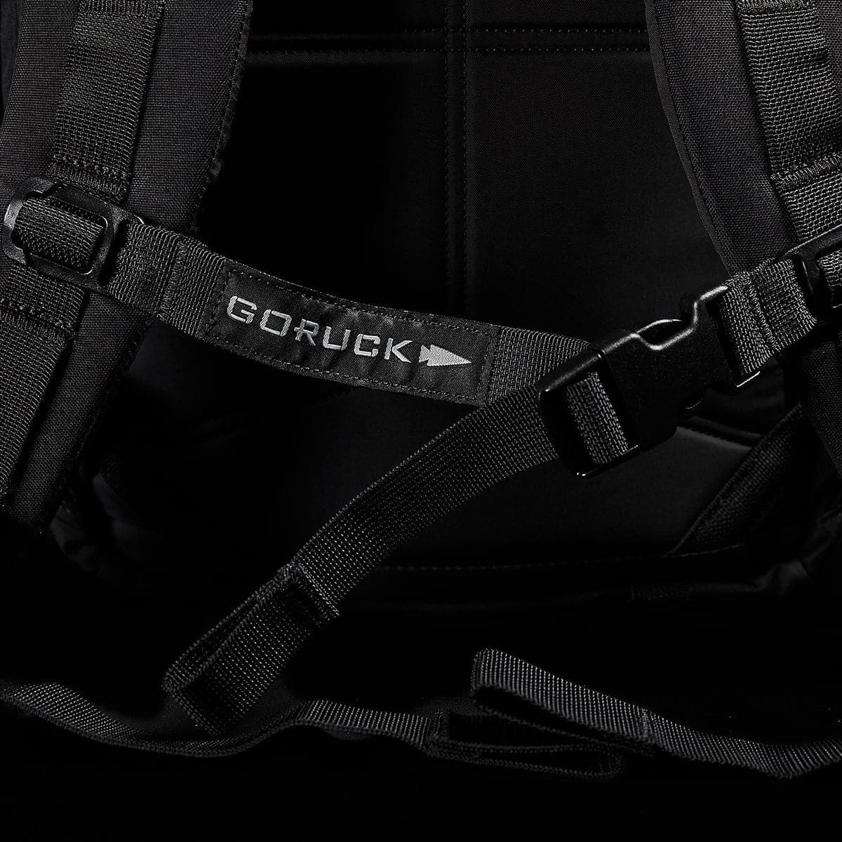 Close-up of a black M23 - Cordura rucksack showcasing the distinct GORUCK strap and buckle. The image emphasizes its modern, tactical design featuring durable materials, reinforced stitching, and adjustable straps set against a dark background.