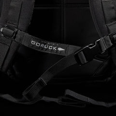 Close-up of a black M23 - Cordura rucksack showcasing the distinct GORUCK strap and buckle. The image emphasizes its modern, tactical design featuring durable materials, reinforced stitching, and adjustable straps set against a dark background.
