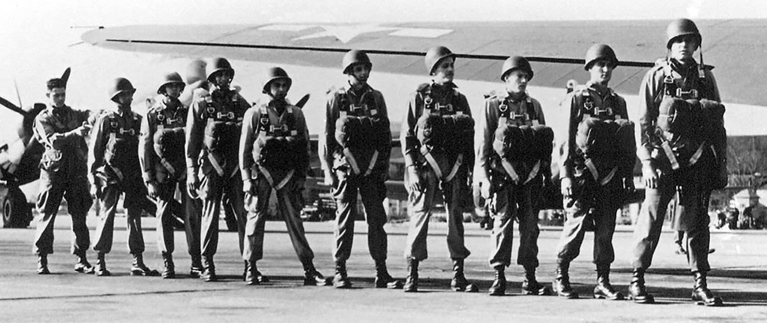 A line of World War II-era paratroopers stands in front of a military aircraft on an airfield.