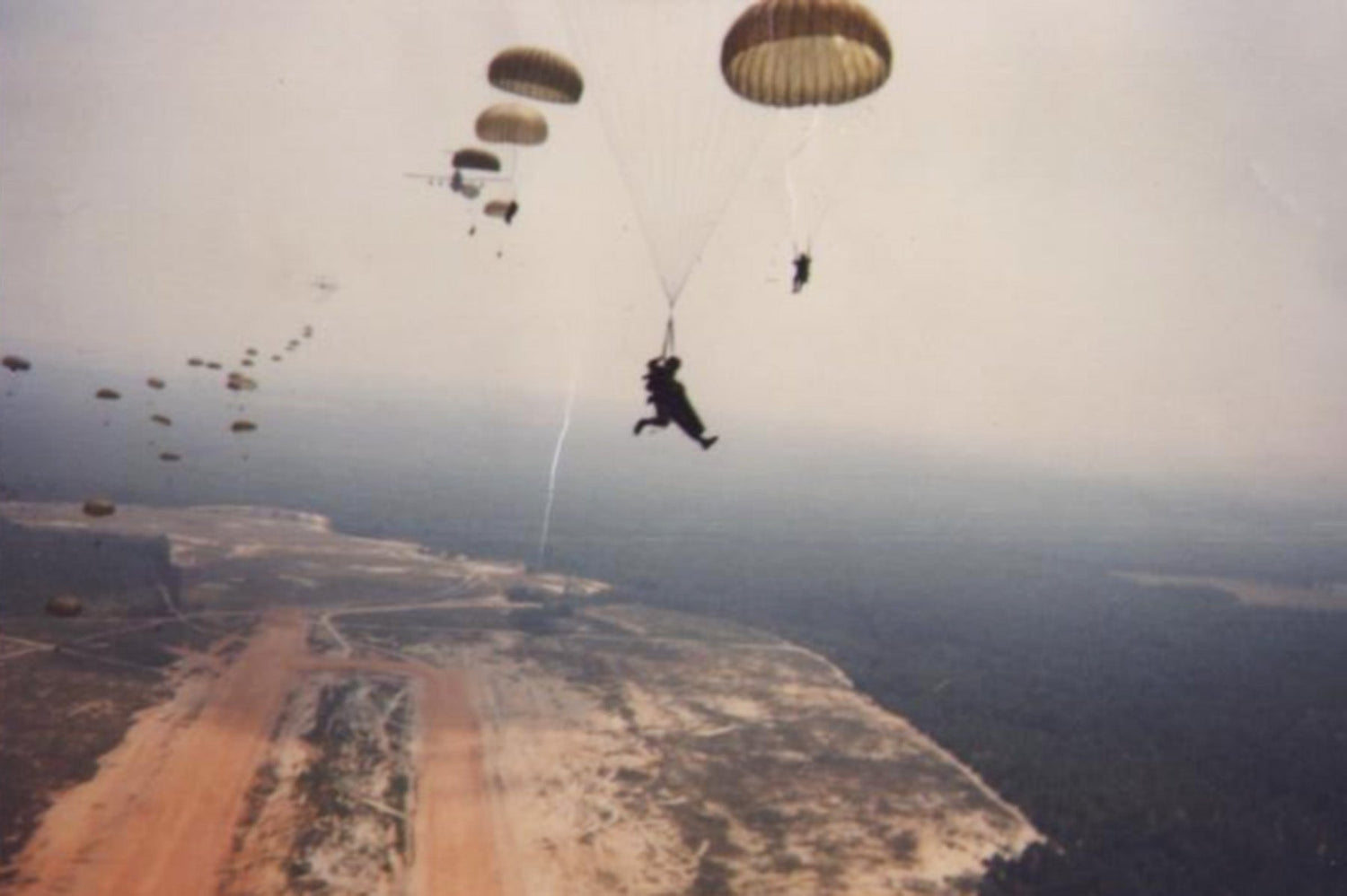Paratroopers descending with parachutes above a wide, open landscape with a dirt clearing below.