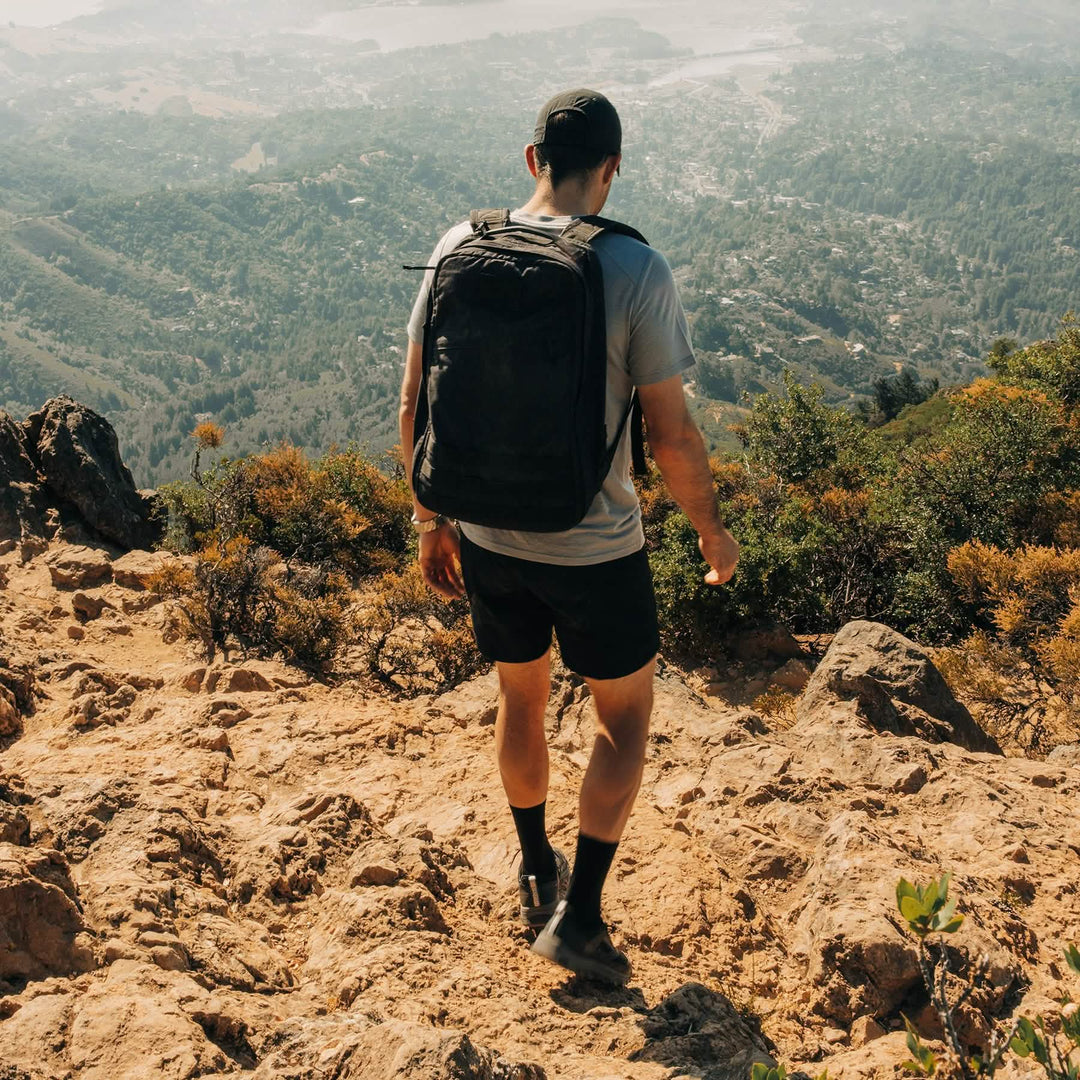 A man with a GORUCK Mackall backpack and Black + Forged Iron trail shoes, designed with a triple compound outsole for extra grip, hiking downhill on a rocky mountain trail with a scenic valley below.
