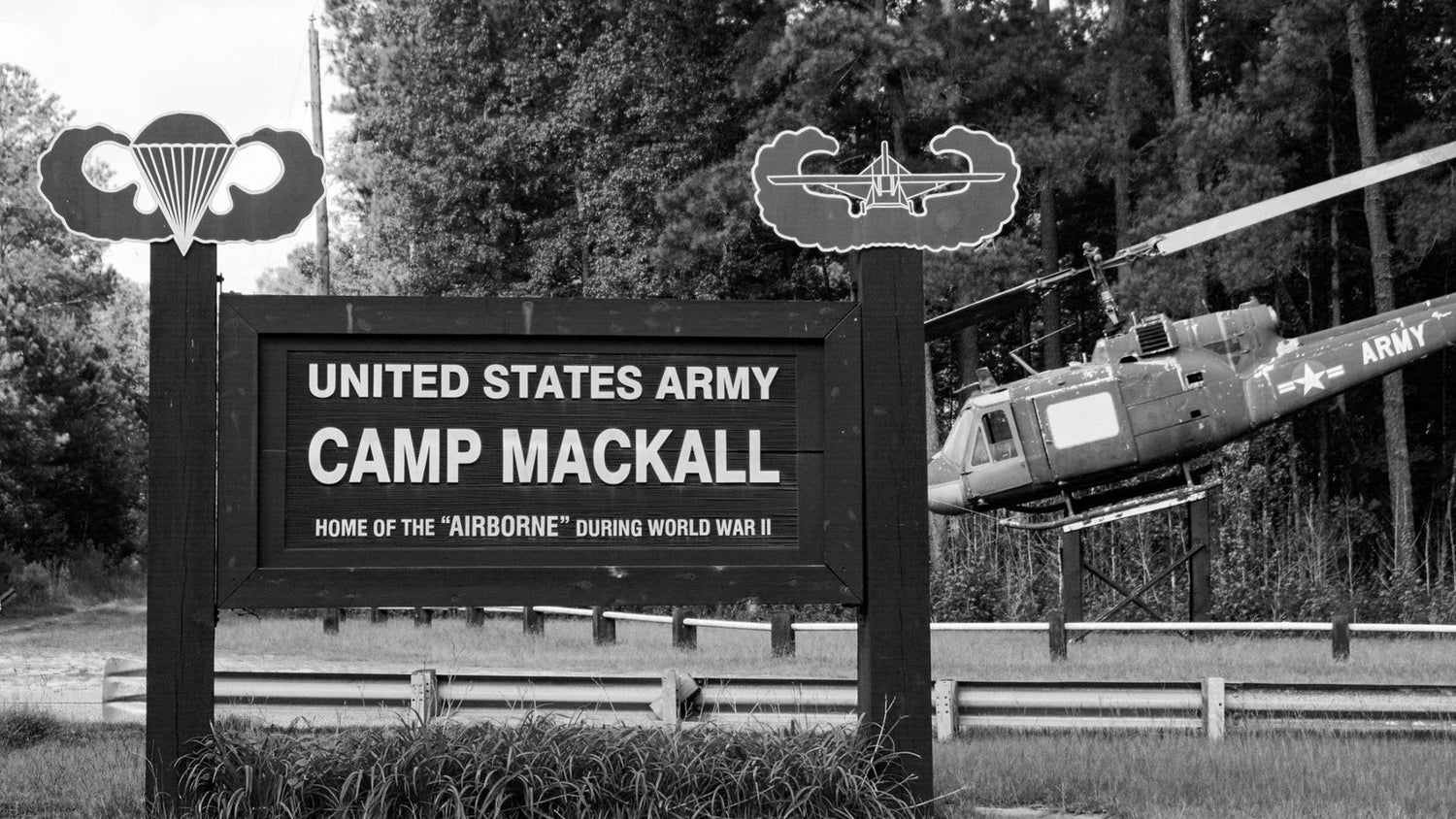 Black and white photo of Camp Mackall entrance sign with a military helicopter in the background.