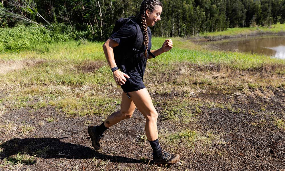 Person with braided hair and a backpack hiking on a dirt path by grassy and wooded areas near water.