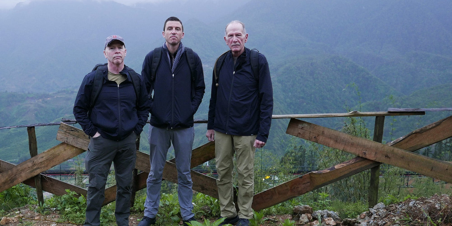 Three men in outdoor clothing stand in front of a wooden fence with misty mountains in the background.