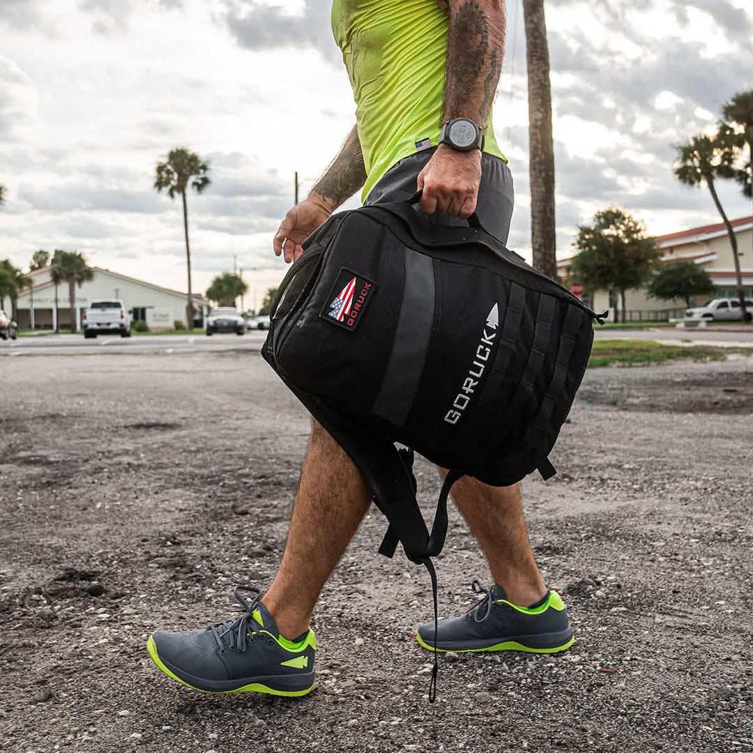 A man in GORUCK Ballistic Trainers – Wolf Grey + Acid Lime with acid lime reflective spearhead walks outdoors, carrying a black backpack made from durable CORDURA® Ballistic Nylon.