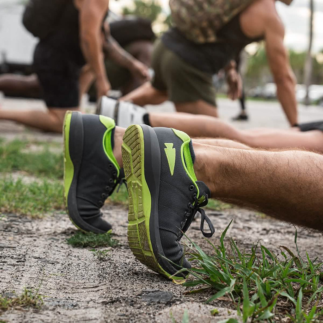 People stretching outdoors in GORUCK Ballistic Trainers - Wolf Grey + Acid Lime W / Acid Lime Reflective Spearhead, featuring a spearhead logo—perfect for those seeking functional fitness footwear.
