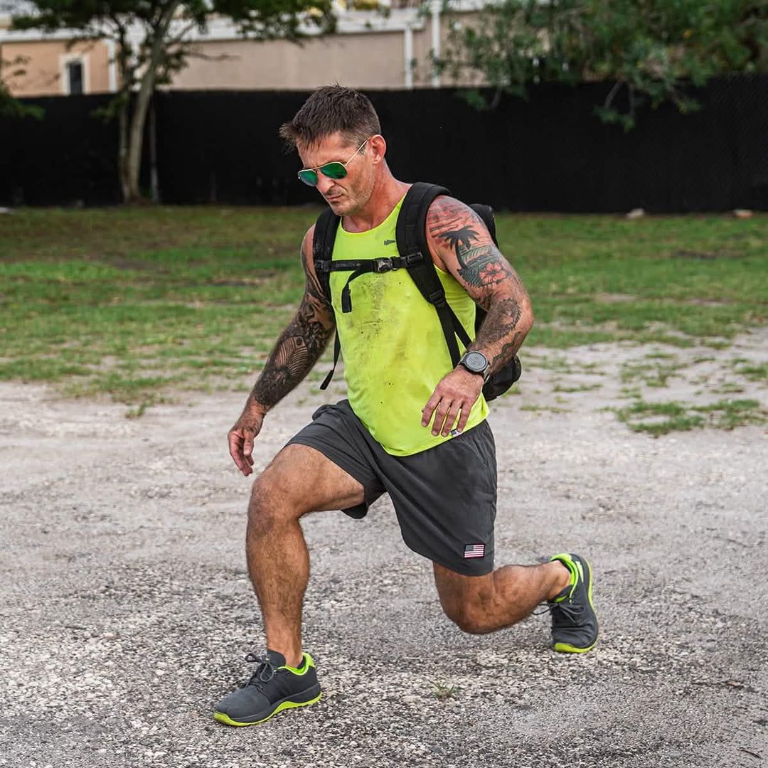 A tattooed man in sunglasses lunges outdoors, sporting a backpack, athletic wear, and GORUCK Ballistic Trainers - Wolf Grey + Acid Lime W / Acid Lime Reflective Spearhead for enhanced support.