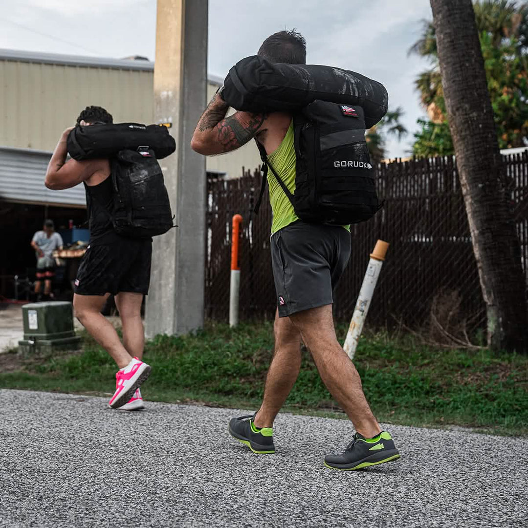 Two men, each carrying heavy sandbags on their shoulders, walk outdoors on a gravel path, showcasing the resilience and stability of GORUCK Ballistic Trainers in Wolf Grey + Acid Lime W / Acid Lime Reflective Spearhead.