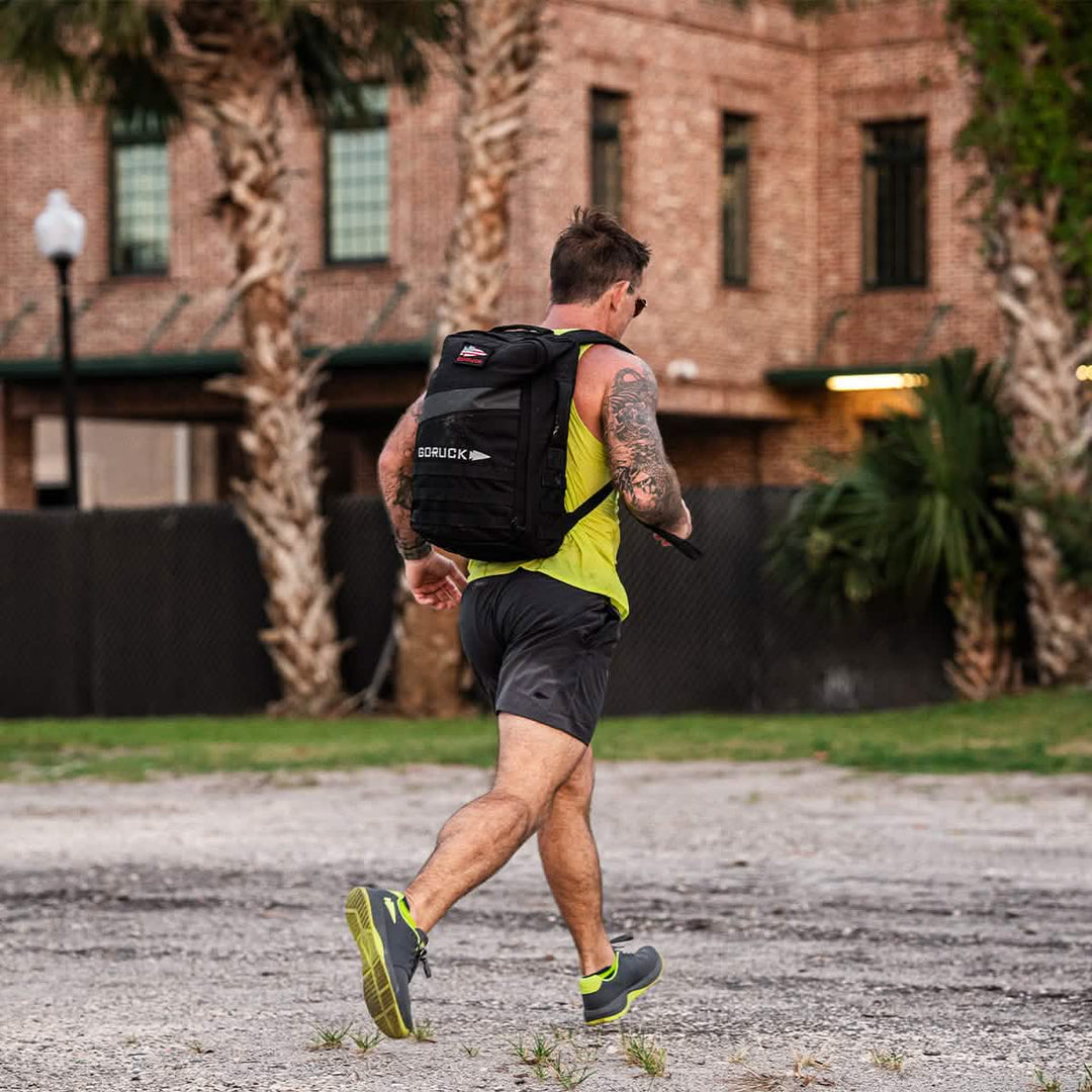 A man in athletic gear jogs outdoors with a black backpack, wearing GORUCK Ballistic Trainers - Wolf Grey + Acid Lime W / Acid Lime Reflective Spearhead. Palm trees and a brick building set the scene for testing his new functional fitness shoes.