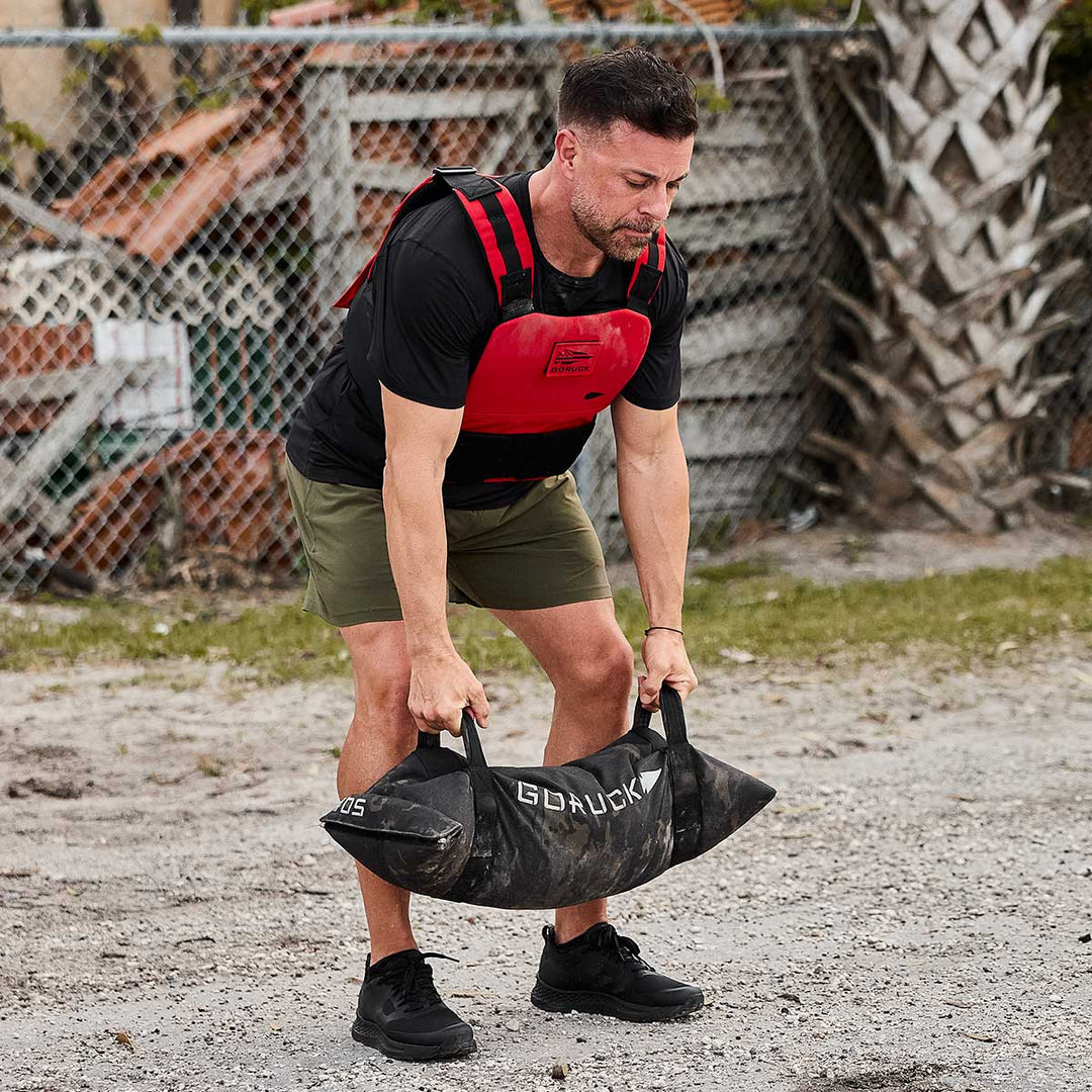 Wearing the Rough Runner - Blackout running shoes from GORUCK, a man dressed in a black shirt and green shorts lifts a sandbag outdoors near a fence. His weighted vest aids his training while the shoes' Gradient Density EVA midsole offers support, and the road-to-trail outsole ensures he's prepared for any terrain.