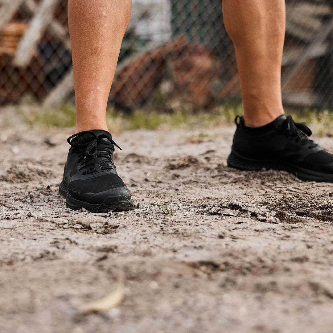 Close-up of a person wearing GORUCK's Rough Runner - Blackout sneakers, featuring a road-to-trail outsole, standing on a dirt path outdoors.