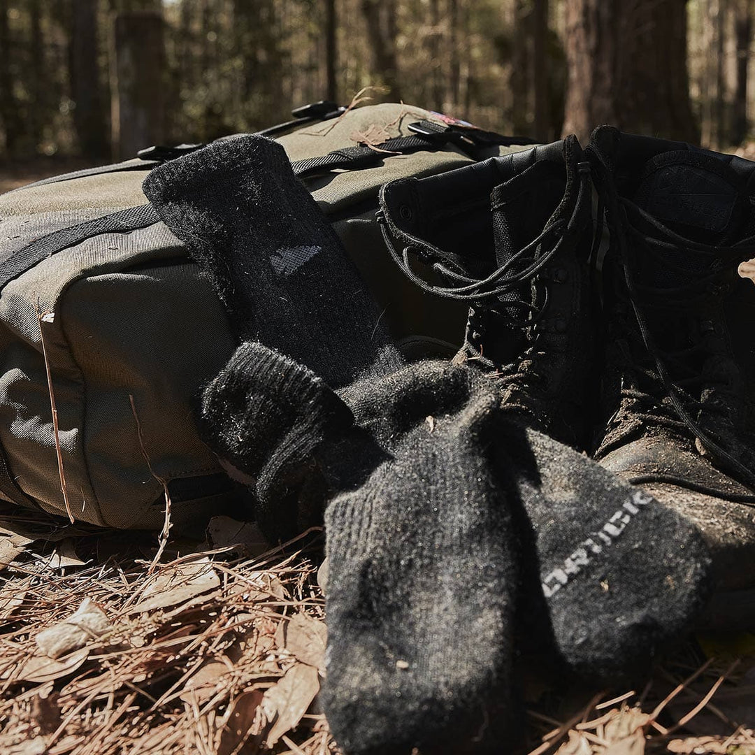 GORUCK's Merino Challenge Socks, a pair of sturdy hiking boots, and a reliable backpack are nestled on the forest floor covered in pine needles, all set for adventure.