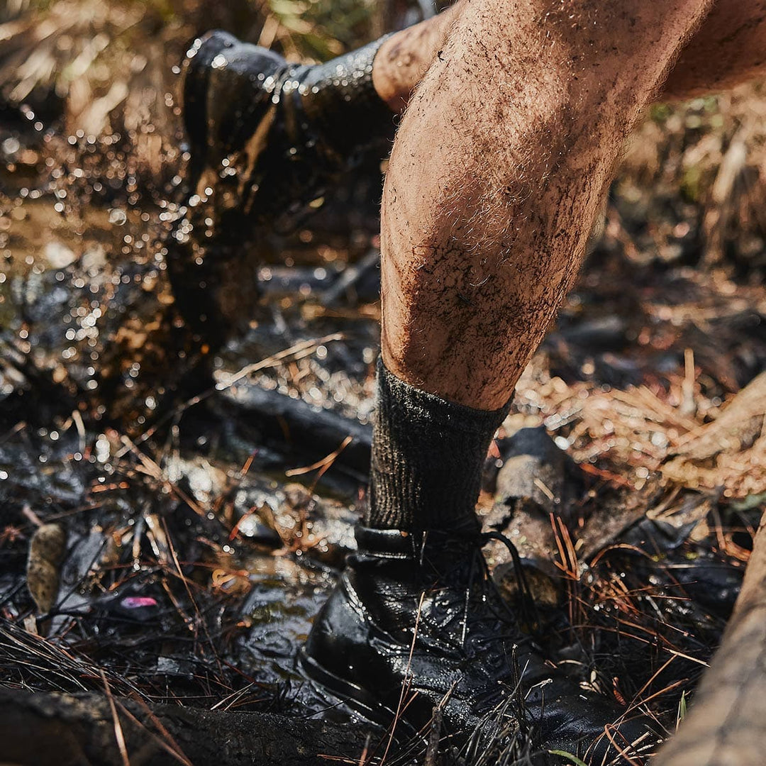 Muddy shoes and legs wading through a murky, waterlogged path in a natural setting, with GORUCK's Merino Challenge Socks providing warmth and comfort every step of the way.