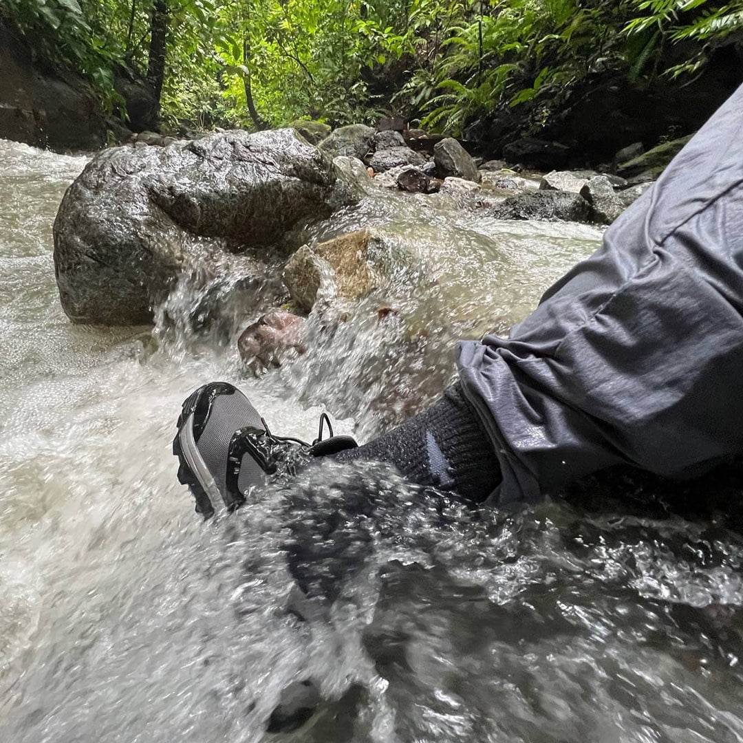 A person in waterproof shoes and Merino Challenge Socks from GORUCK crosses a rocky stream in a forest, confident in their lifetime guarantee.