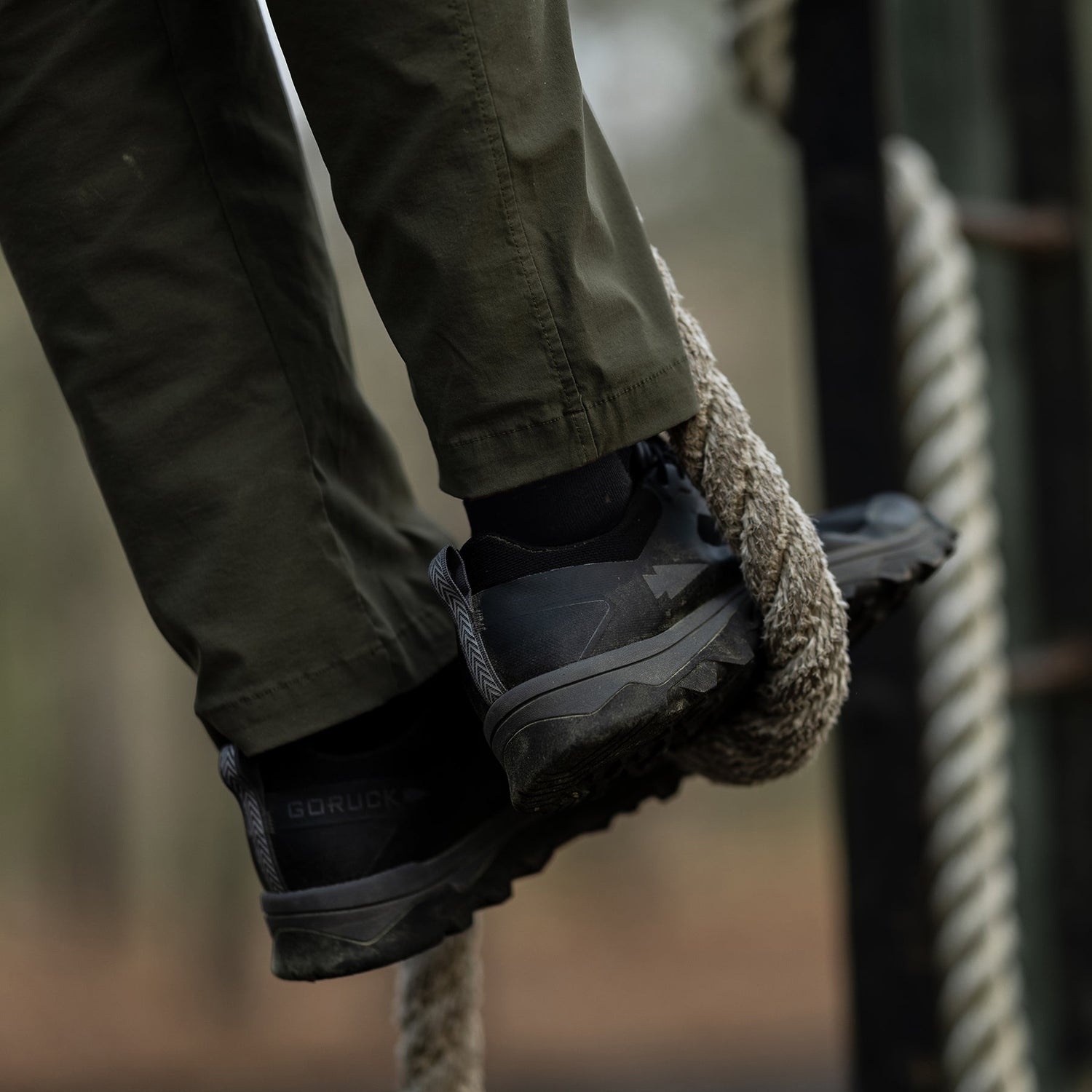 Person wearing black shoes and green pants climbing a thick rope outdoors.