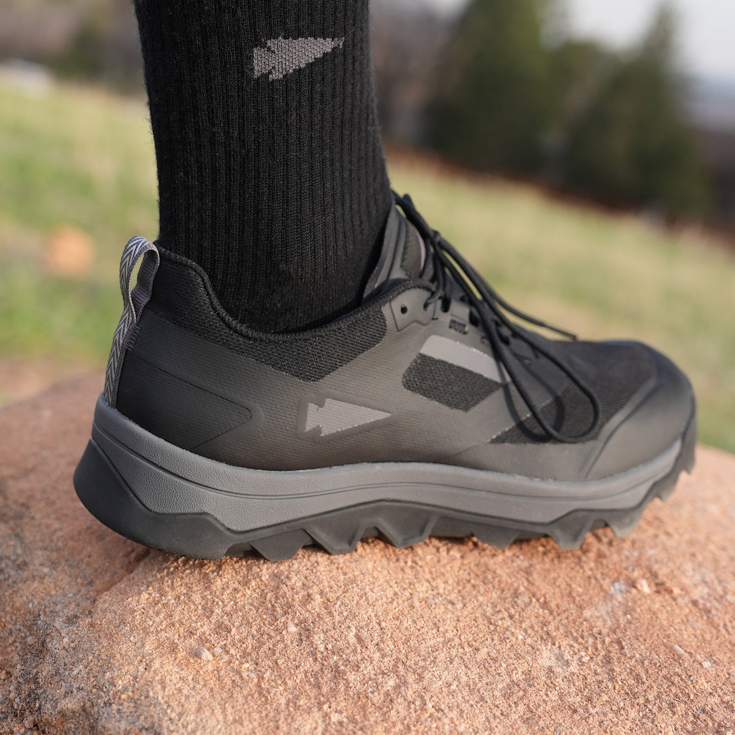 A close-up of a black trail running shoe and sock on a person's foot, standing on a rock outdoors.